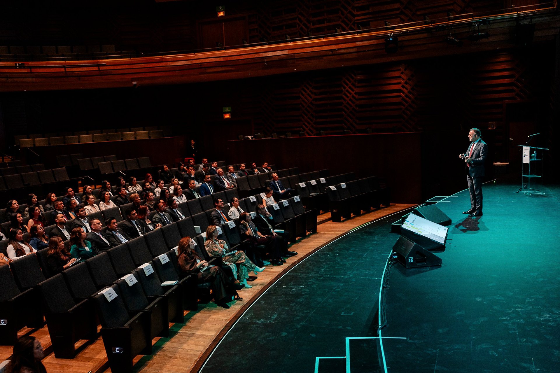 Speaker presenting on stage under turquoise lighting to a seated audience in a modern auditorium