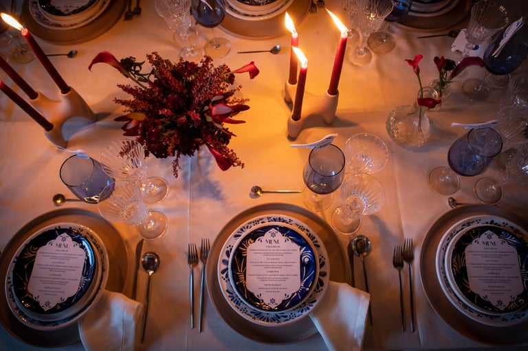Elegant dinner table setting with three place settings, decorative plates, candles, and a red floral centerpiece under warm lighting