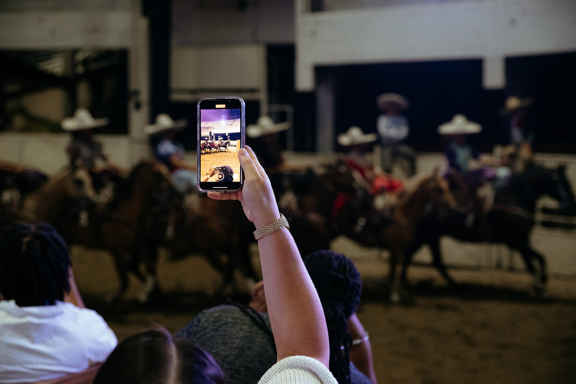 Person holding up smartphone to record horse riding event in indoor arena with crowd in background