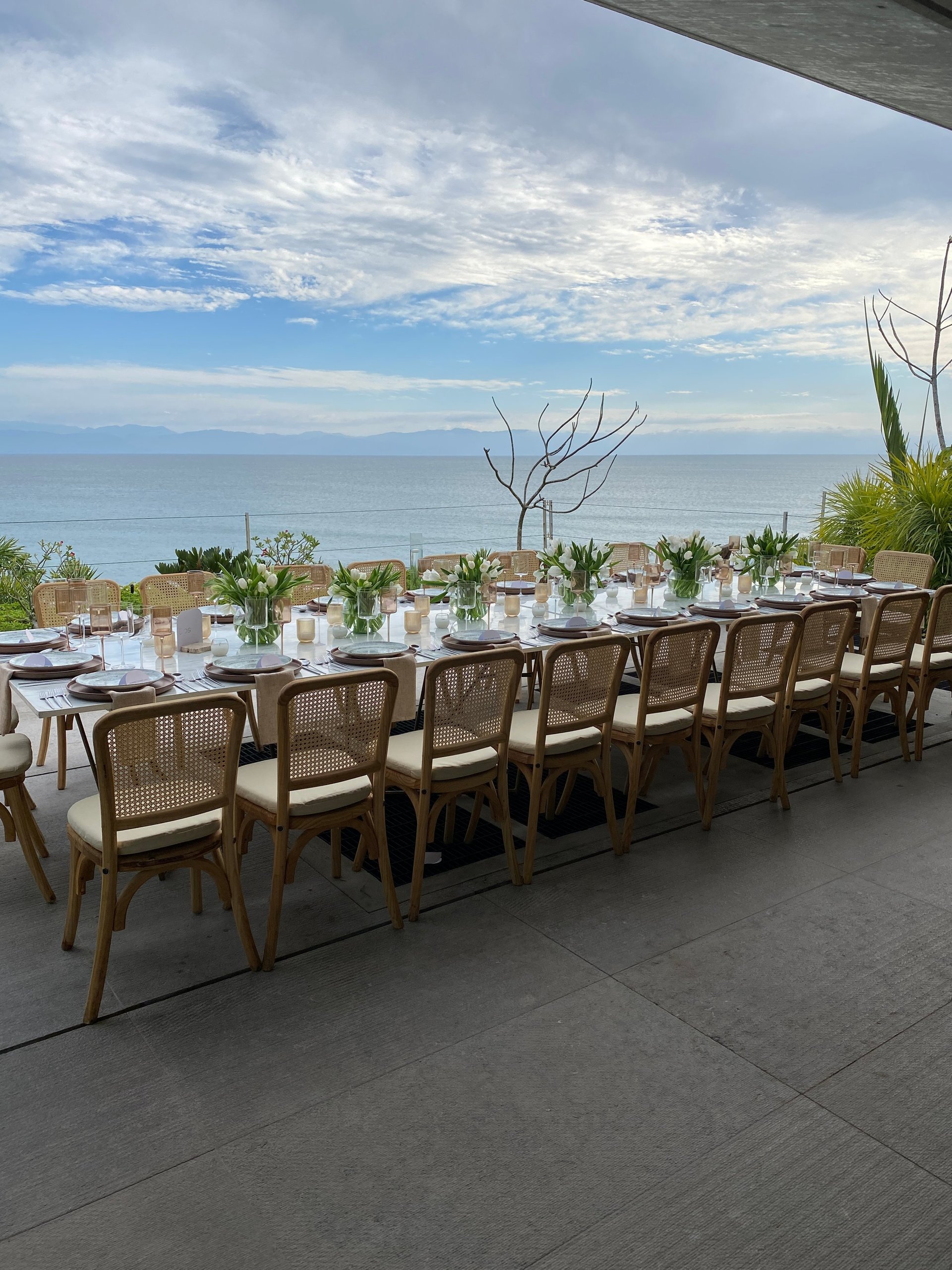 Long outdoor dining table with wooden chairs overlooking the ocean, decorated with candles and floral arrangements
