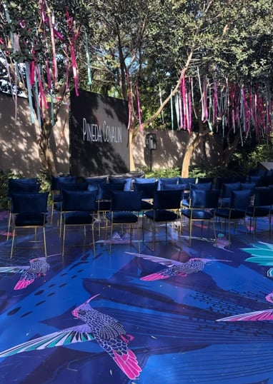 Outdoor event space with blue chairs arranged in a circle, decorated with pink hanging ribbons and a blue floor featuring fish designs under trees