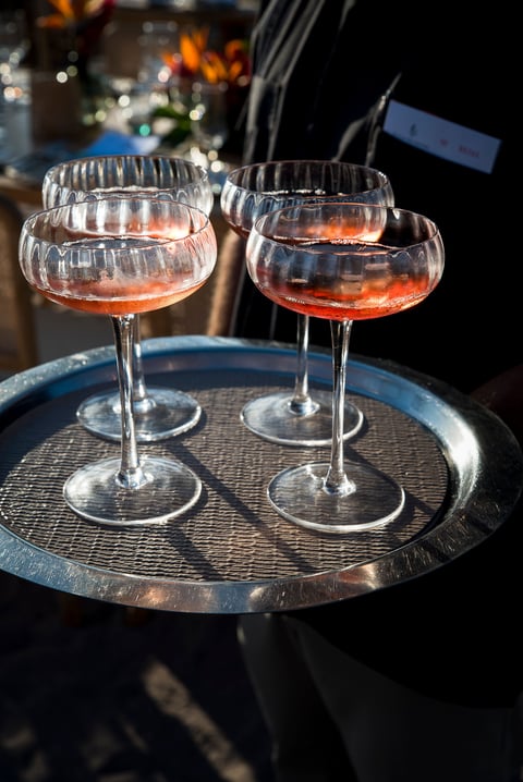 Four coupe glasses filled with rose-colored cocktails on a circular metal tray at an evening gathering