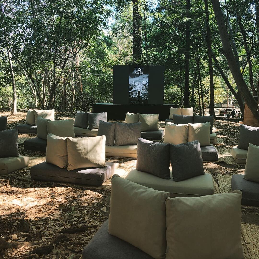 Outdoor forest gathering space with green and beige floor cushions arranged in a circle facing a black screen mounted on a wooden structure