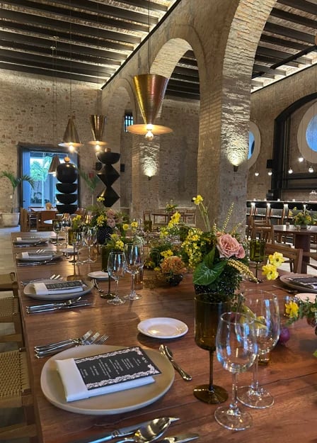 Elegant dining room with wooden table, place settings, and floral centerpieces under arched brick architecture with pendant lighting