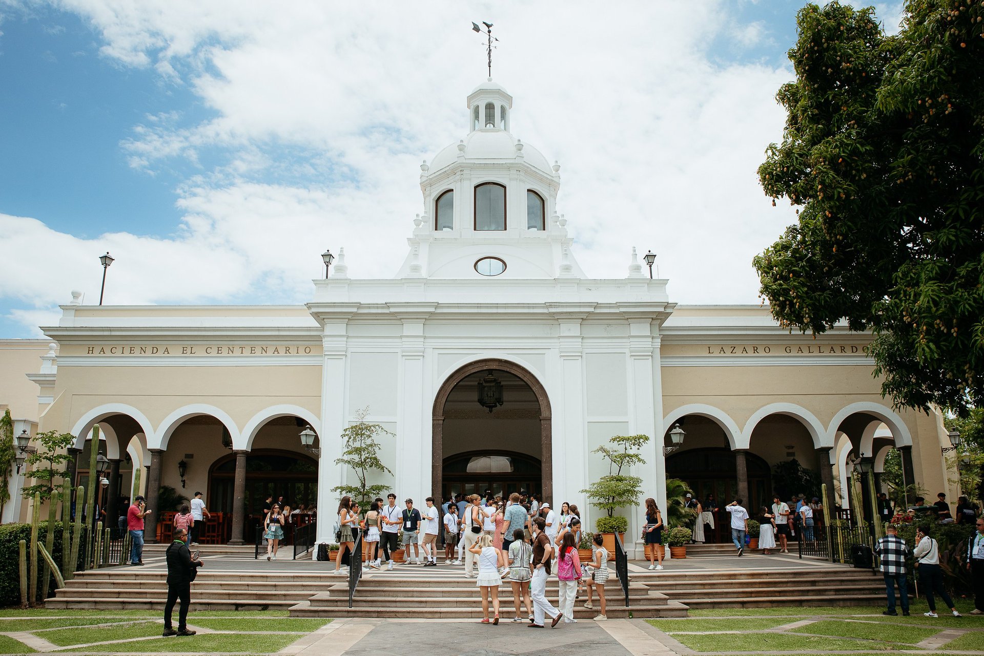 White historic building with dome and arched entrance, visitors gathering on front steps and plaza on sunny day