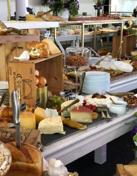 Bakery display counter with assorted breads, pastries, cheeses, fresh vegetables, and white plates arranged on tiered shelving