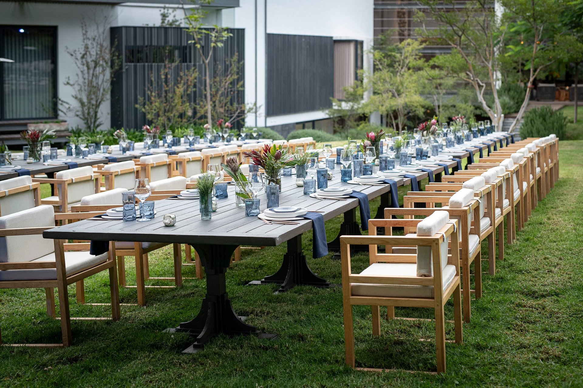 Long outdoor dining setup with wooden chairs and black tables decorated with blue glassware and flowers in a garden