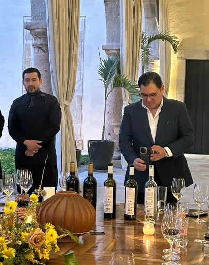 Two men in business attire at a wine tasting event with bottles displayed on a wooden table and fall floral arrangements