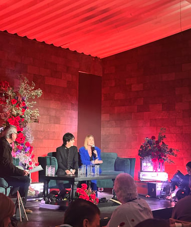 Two people sitting on a blue sofa on stage with vibrant red and pink floral arrangements and neon lighting in the background