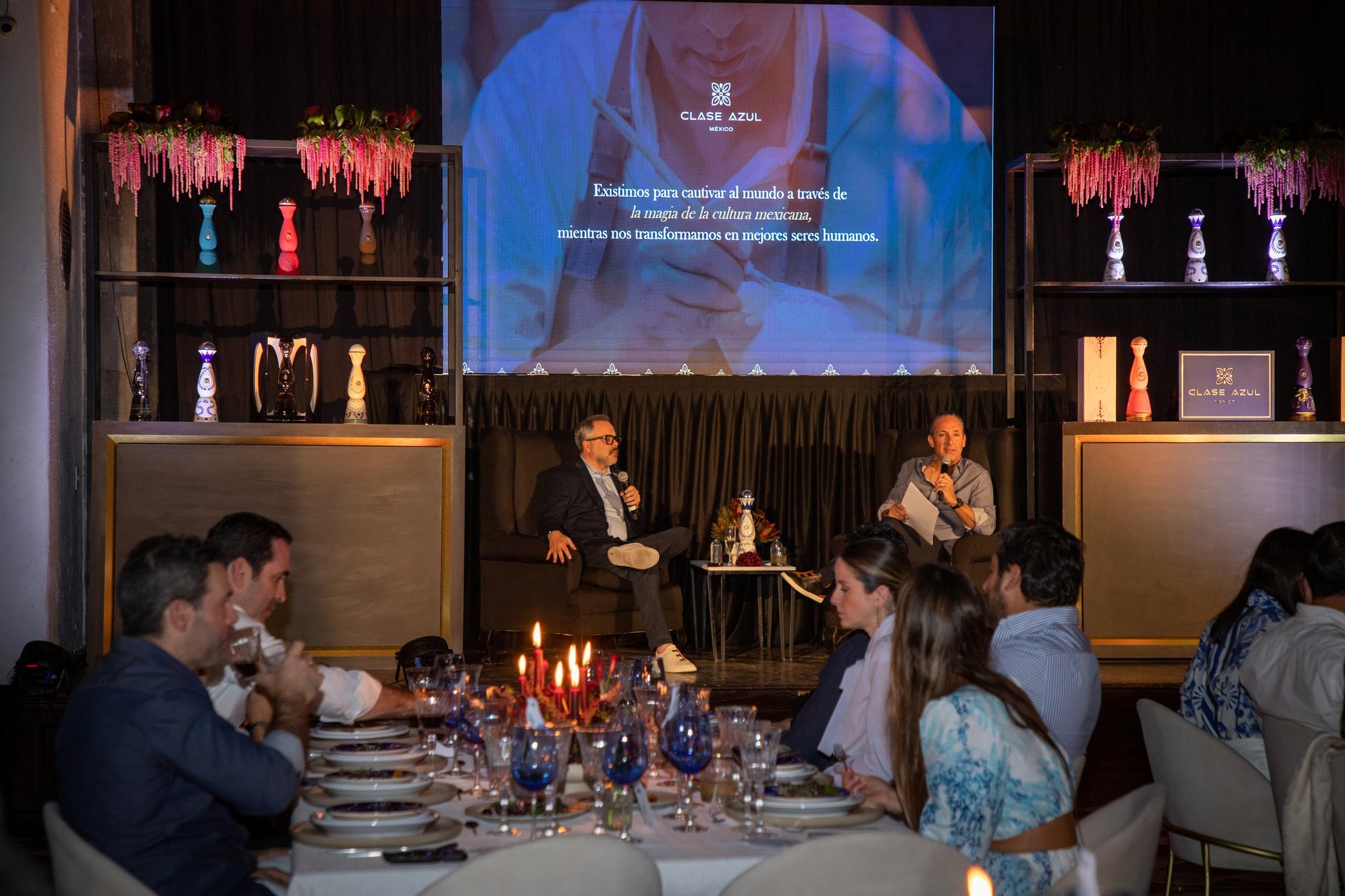Professional conference setting with attendees seated at a dining table while two speakers present on stage beneath a large projection screen